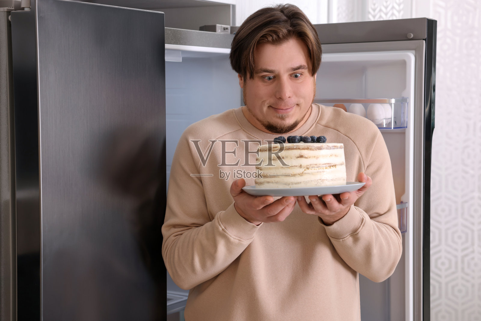 快乐的胖男人在厨房开着的冰箱旁边放着蛋糕照片摄影图片