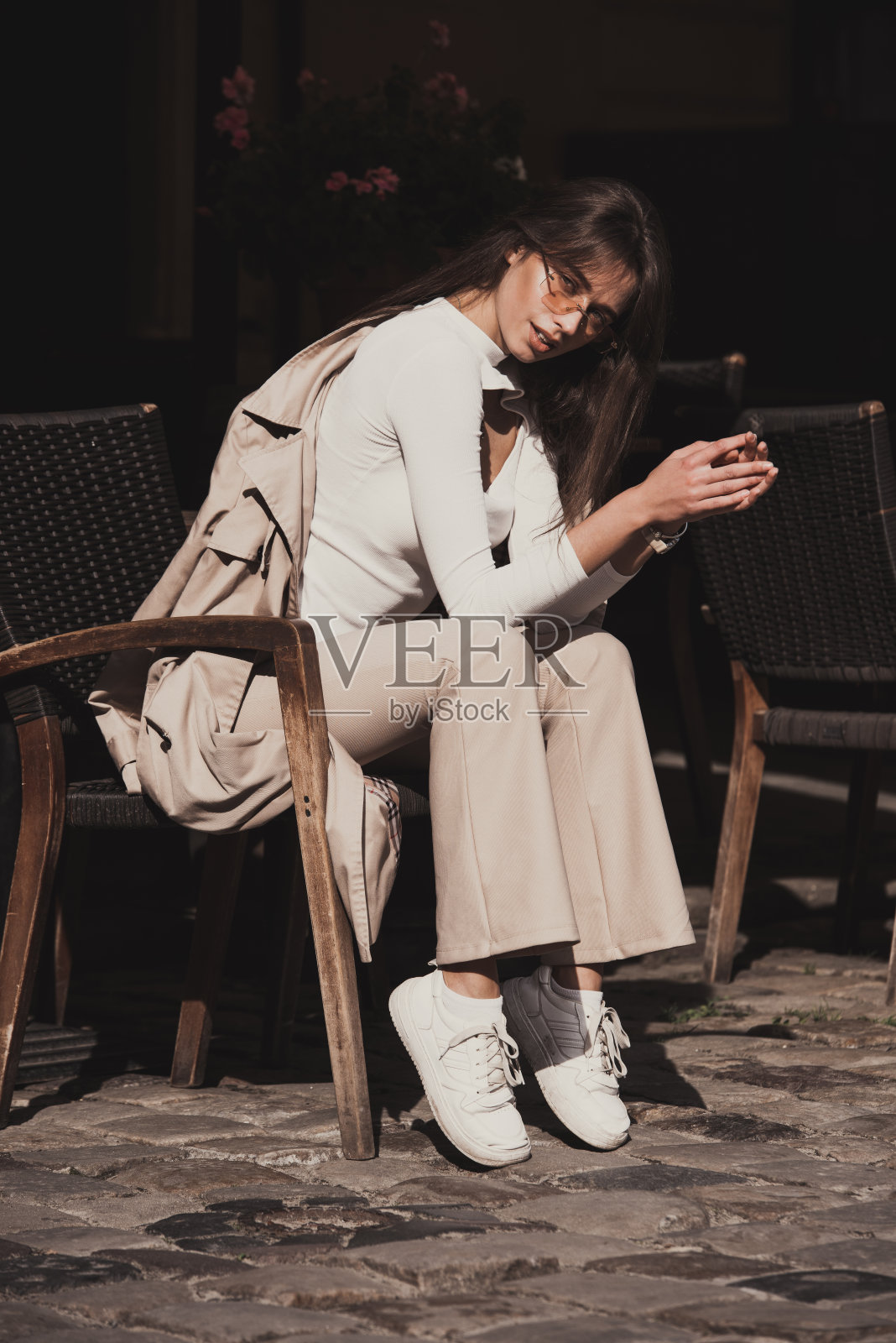 美丽的褐发女孩穿着白色衬衫，裤子，运动鞋与手提包和驼色外套在手中。时髦时髦的时尚观照片摄影图片