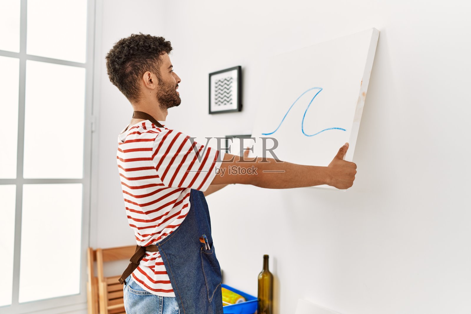 年轻的阿拉伯艺术家微笑着快乐地挂在艺术工作室的墙上画画布。照片摄影图片