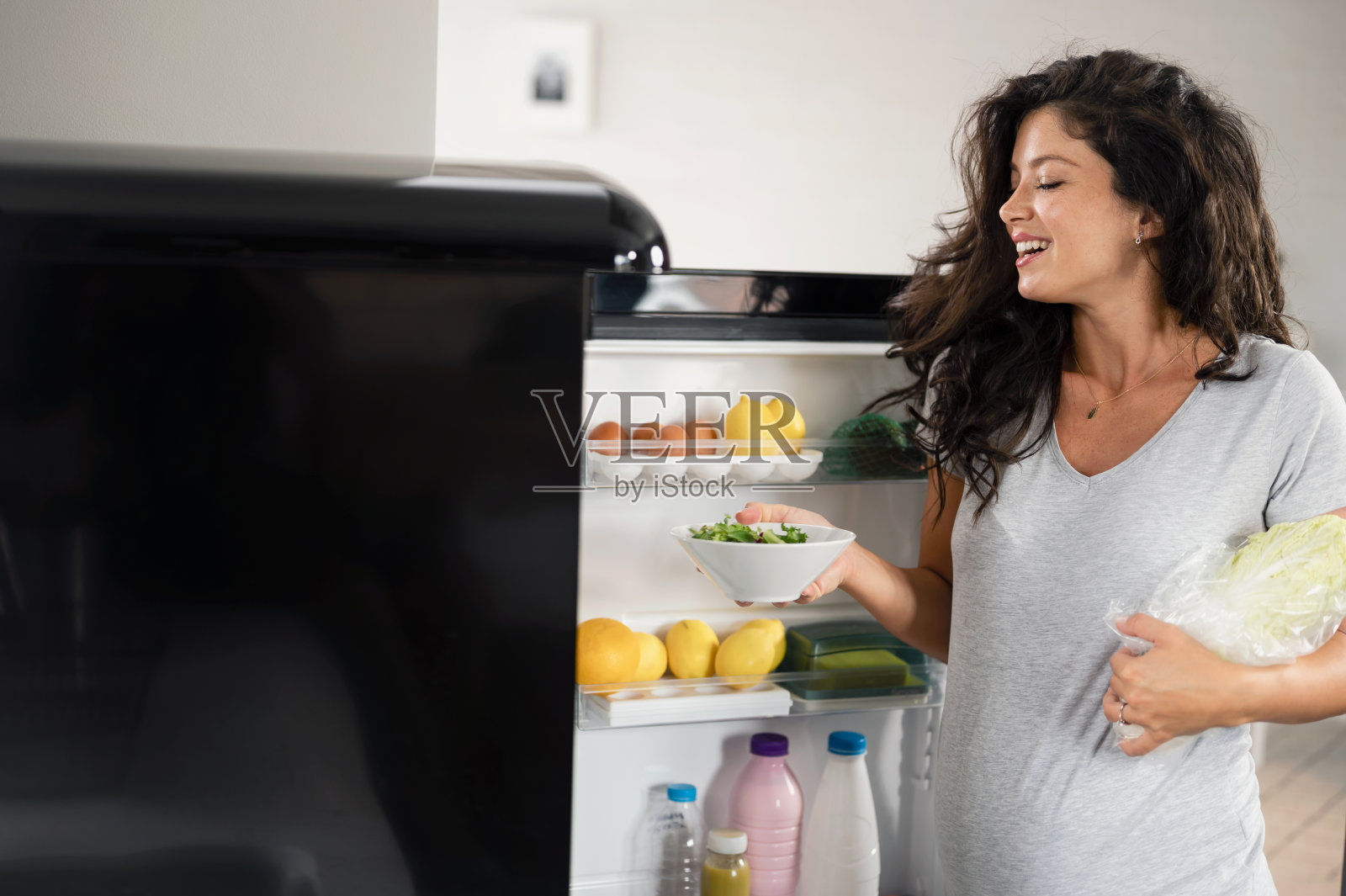 美丽年轻微笑的孕妇从冰箱里拿健康食物。怀孕期间的健康饮食水果、奶制品和谷物。照片摄影图片