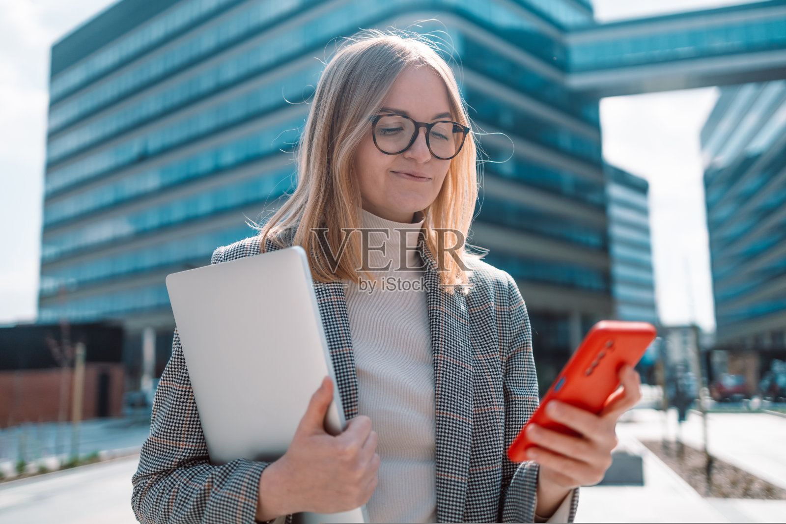 成功的金发女人的肖像使用智能手机，手持数字平板电脑在城市建筑的背景。商务人概念照片摄影图片
