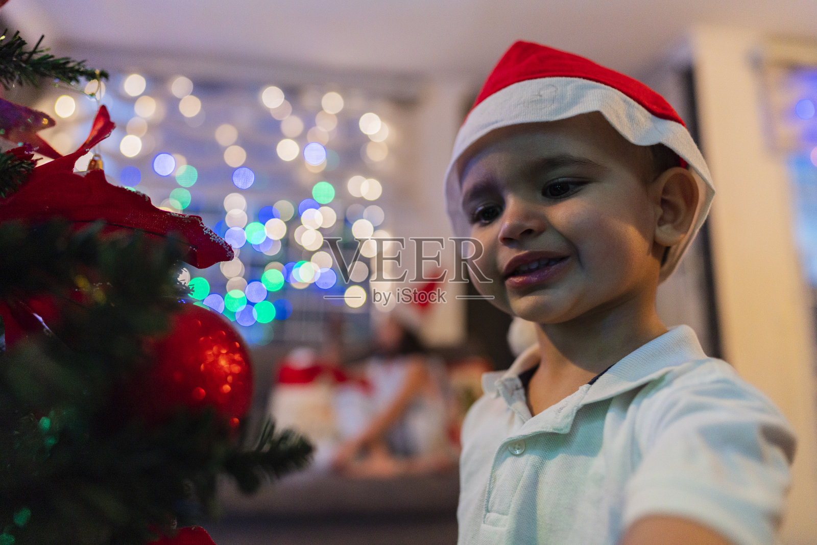 一个拉丁美洲的男孩在他为圣诞节而装饰的房子的客厅里，他正把圣诞球挂在他的树上照片摄影图片