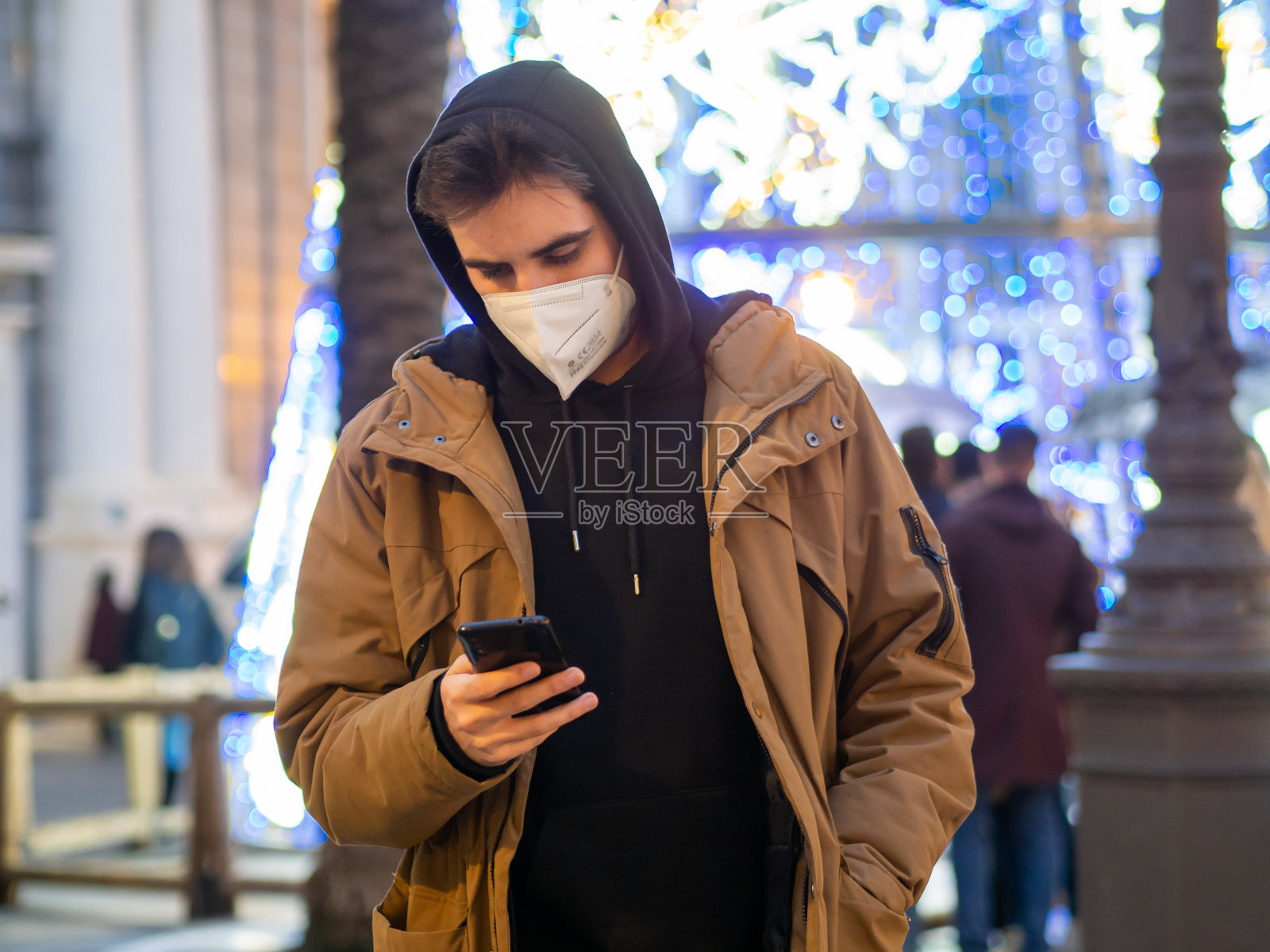 戴着口罩的年轻男性在圣诞节期间看着他的手机，旁边是一棵点亮的圣诞树照片摄影图片