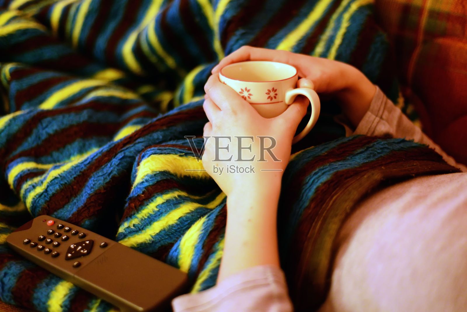 女人坐在家里的毯子下，手里拿着遥控器和茶杯照片摄影图片