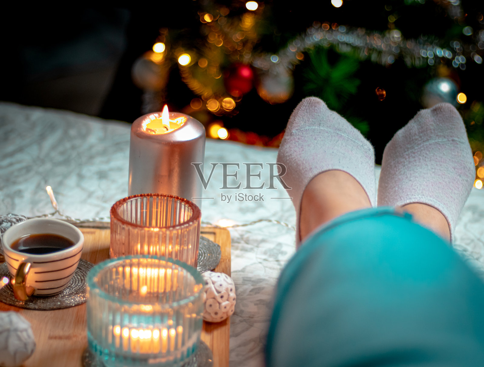 女人穿袜子的腿，一杯卡布奇诺，毯子上的蜡烛和一棵挂满彩灯的圣诞树。家的舒适和舒适的气氛照片摄影图片