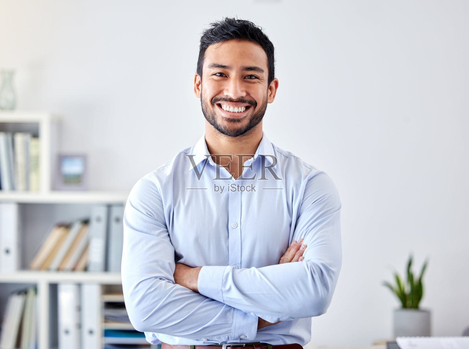 在一家初创公司的现代化办公室里，一个商人面带微笑。快乐的职业经理人或企业家双臂交叉站在他的工作场所。照片摄影图片