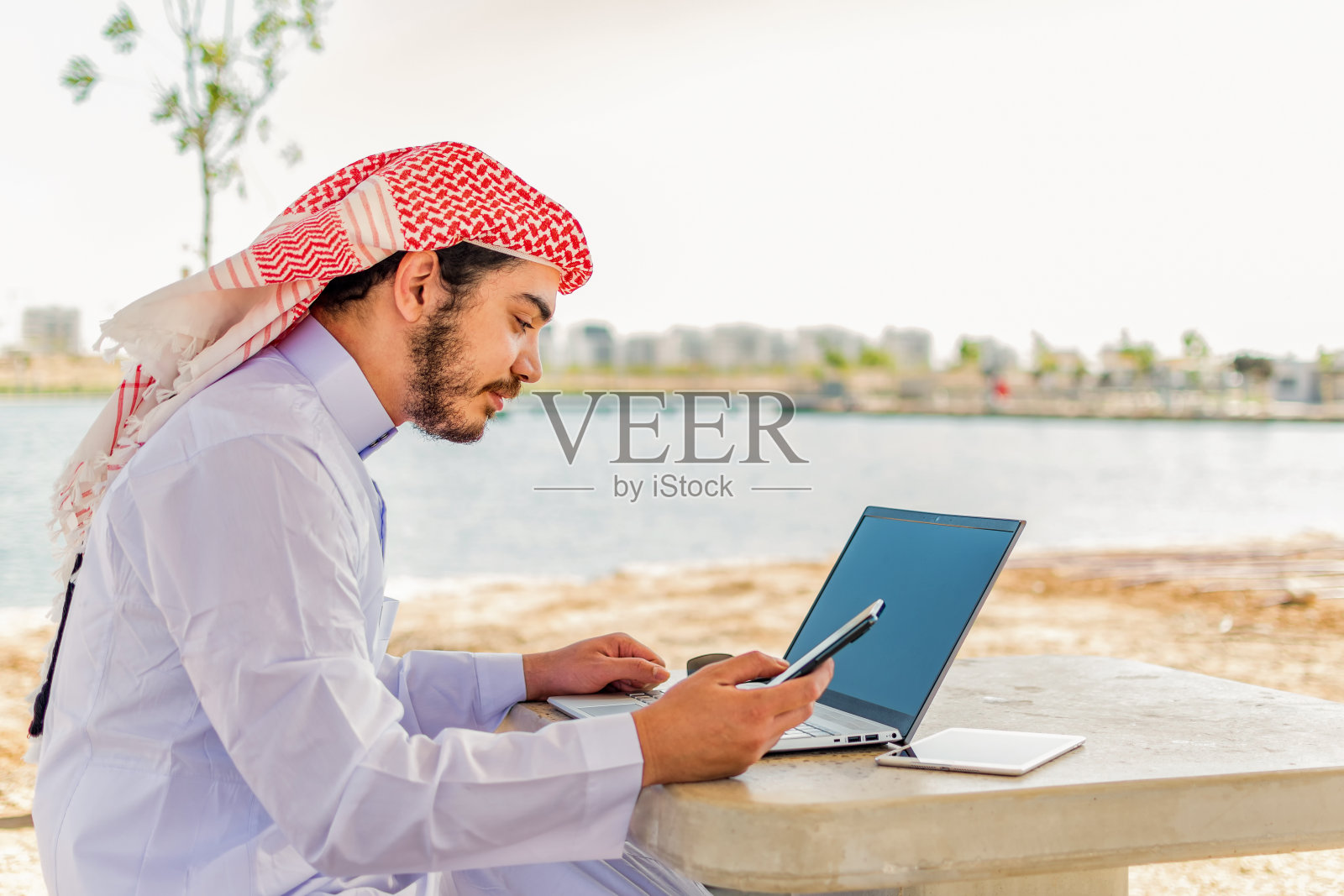 在中东海域，一名阿拉伯男子正在用笔记本电脑和电话工作。照片摄影图片