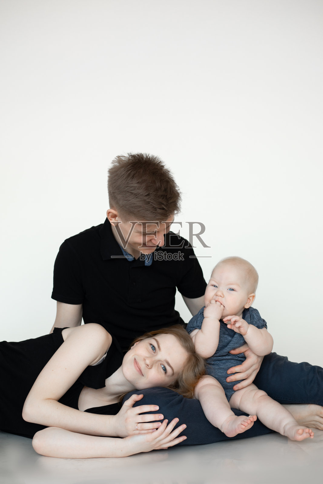 年轻的三口之家在白色的背景下，穿着黑色的衣服。爸爸坐在地板上抱着婴儿舔着婴儿的手，妈妈高兴地躺在丈夫的腿上。全家福，育儿，长子，爱，感情照片摄影图片