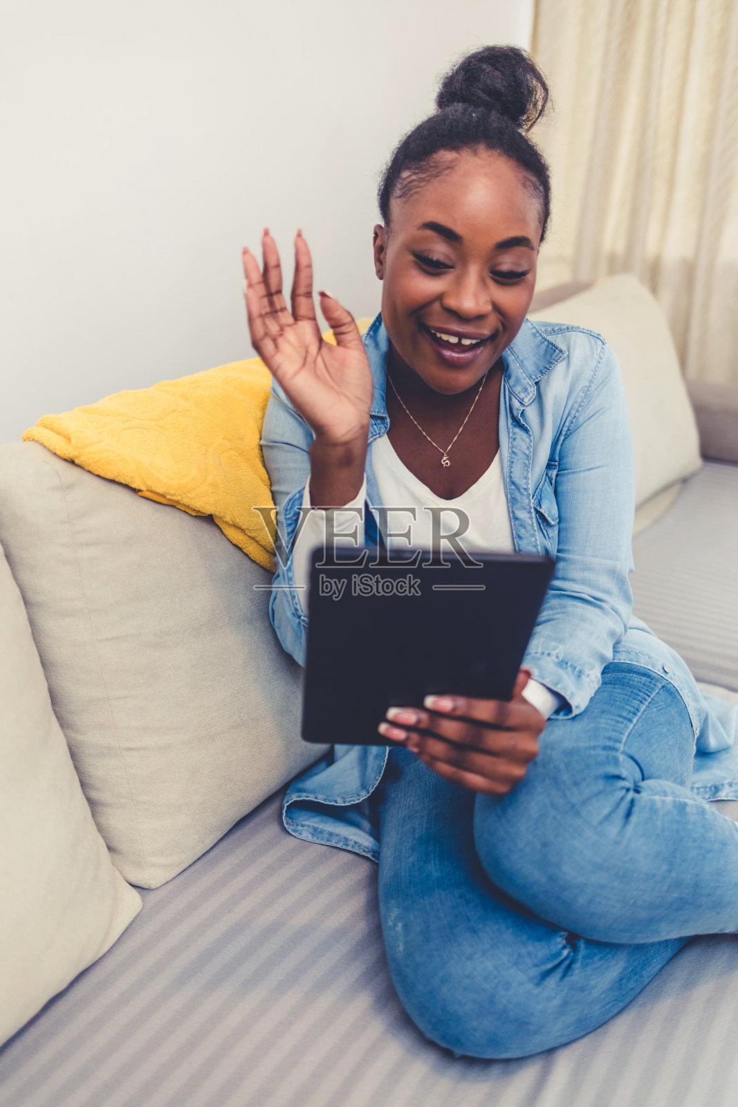 迷人的黑人妇女在家里拿着平板电脑。快乐的女人通过在线视频通话，在家里用笔记本电脑。年轻女子舒适地坐在客厅的沙发上。照片摄影图片