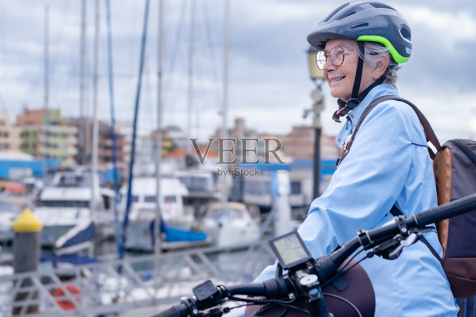 微笑年长的白人妇女戴着自行车头盔看着船停泊在港口，站在她的自行车附近。迷人的女性享受自由和假期照片摄影图片