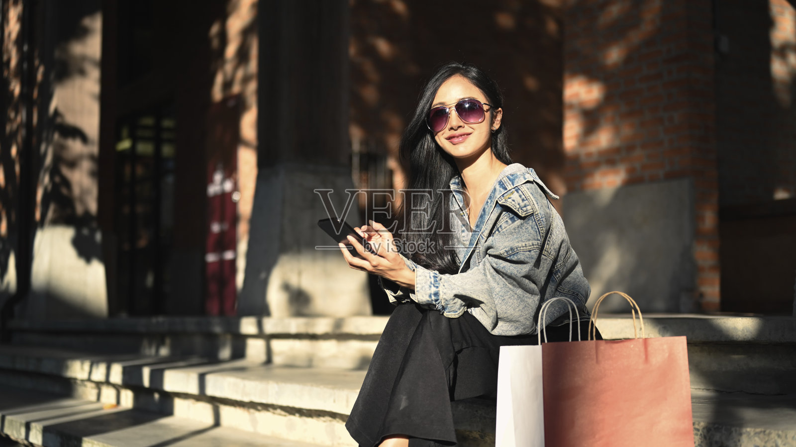 现代千禧一代女性的肖像，穿着时髦的衣服，戴着太阳镜，坐在一个城市购物区的楼梯上。照片摄影图片