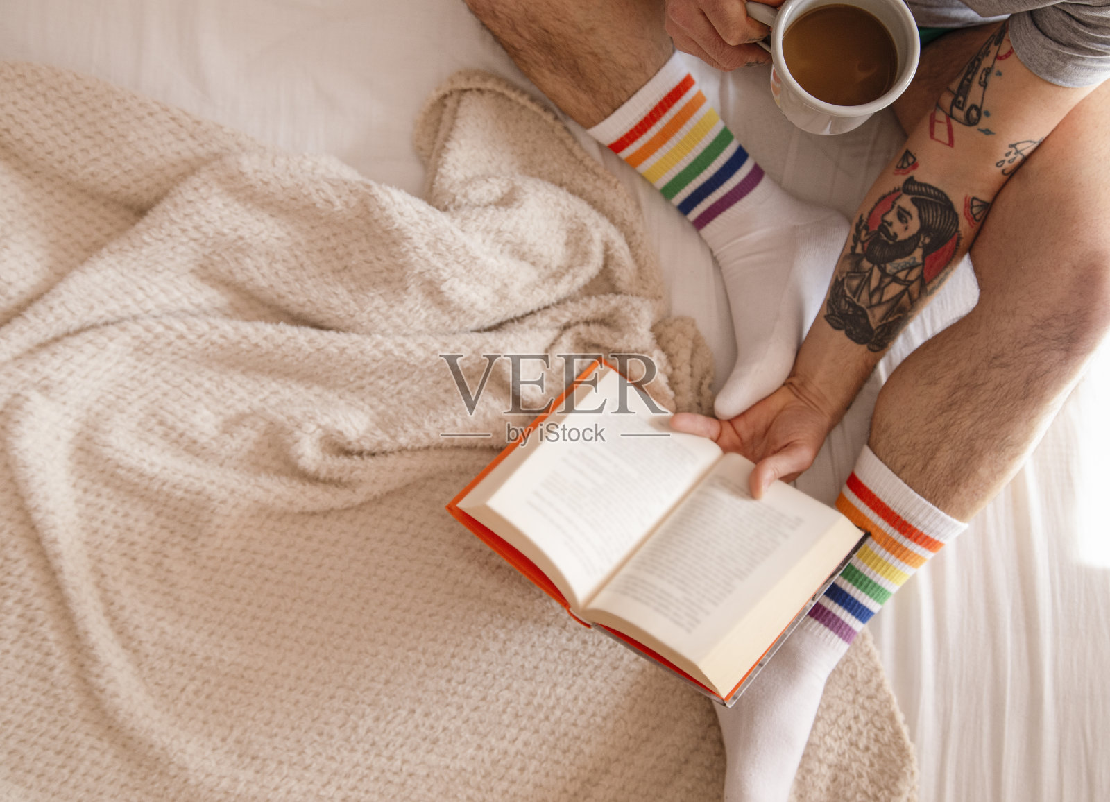 俯视图上，一个穿着彩虹袜的纹身男子早上躺在床上喝咖啡看书照片摄影图片