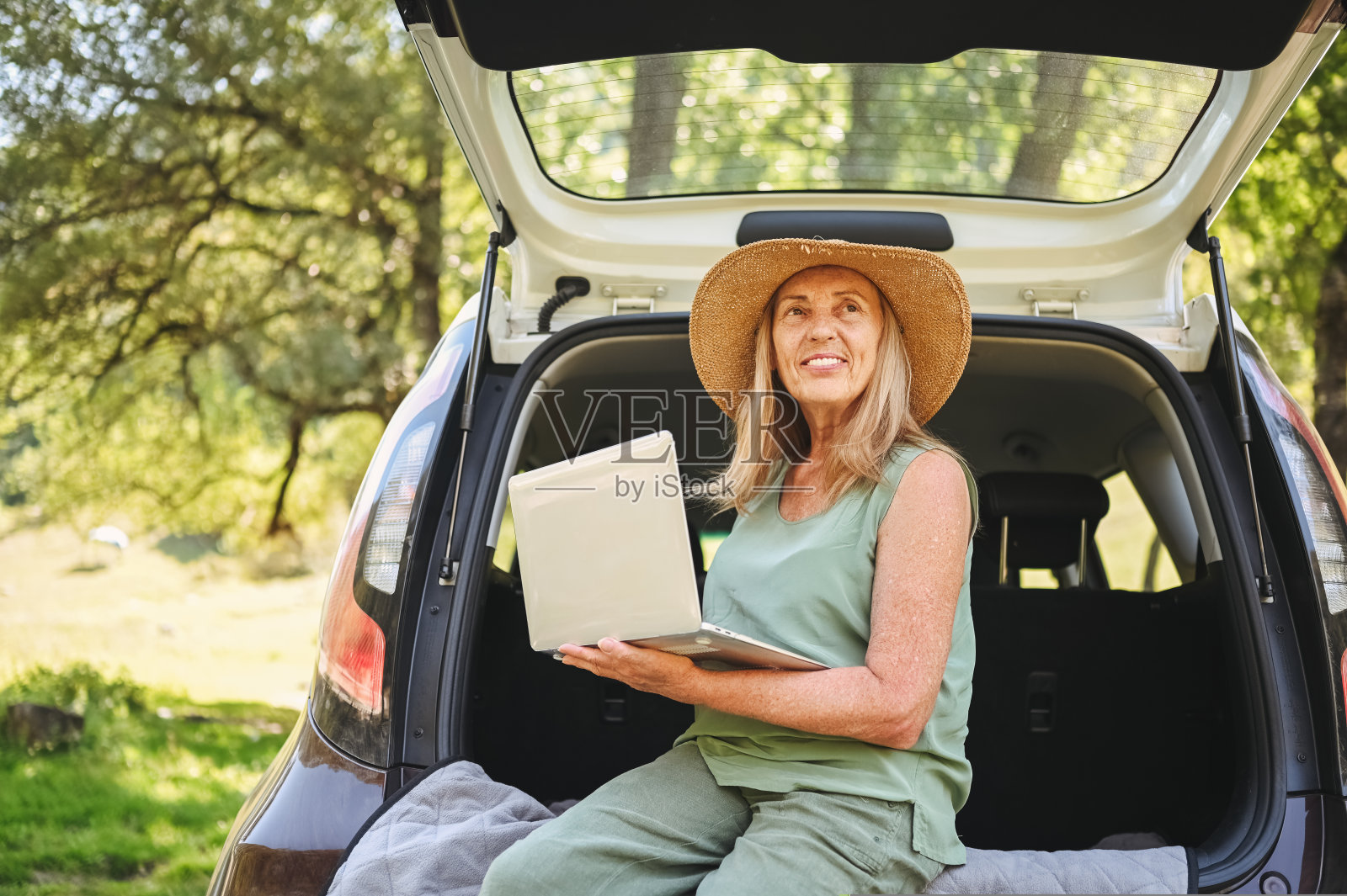 快乐的妇女工作与笔记本电脑从汽车后备箱或在露营者阳光明媚的夏季公园-数字游牧自由生活方式的概念，旅行自由职业者和退休的老年人的假期..照片摄影图片