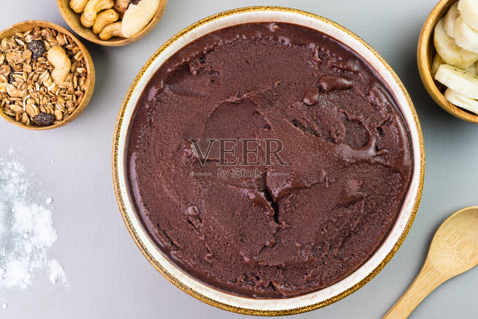 Brazilian açai in a white bowl, additional banana, granola and nuts in small bamboo bowls. Cashew and Pará nuts. Top view. Gray background. Selective focus照片摄影图片