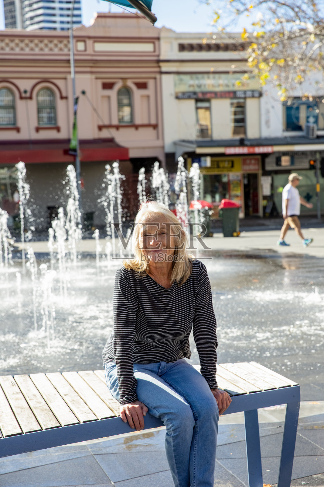 快乐的老年妇女坐在附近的水喷泉在城市，正面视图，背景与复制空间照片摄影图片