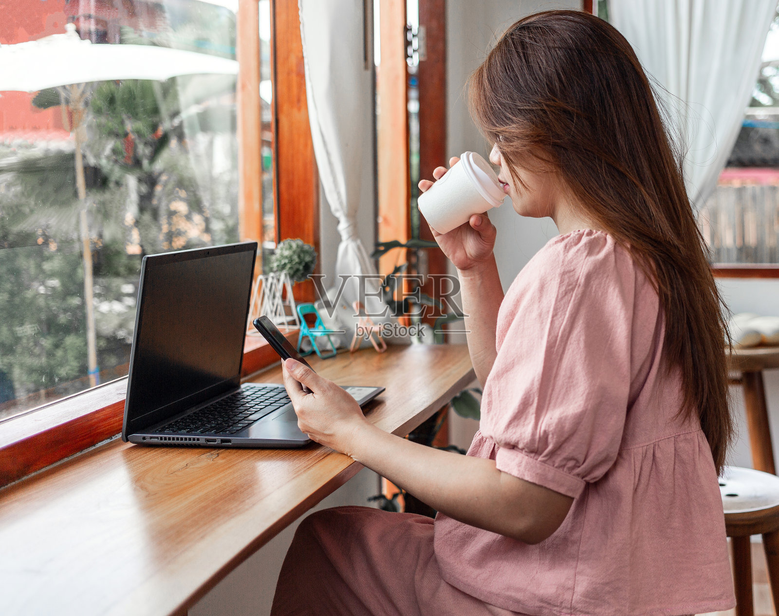 在咖啡厅用智能手机和笔记本电脑喝纸杯咖啡的快乐女性。一头长发的年轻白人女子坐在咖啡店里，忙着玩她的智能手机。照片摄影图片