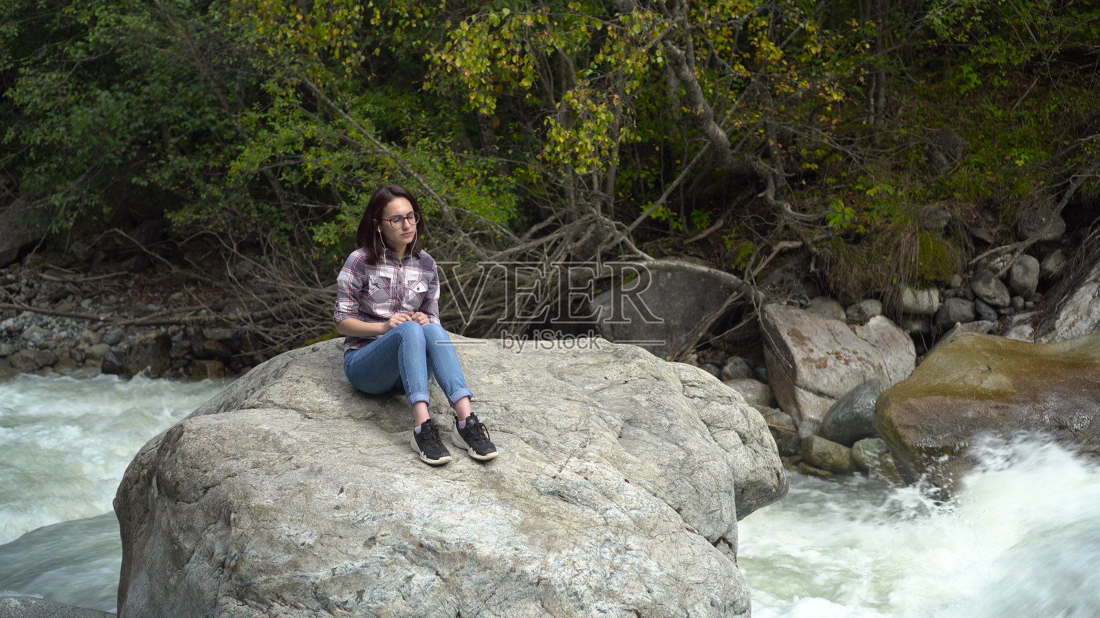 在河边的山上，一位戴眼镜的年轻女子戴着耳机坐在一块石头上。去山区旅行。照片摄影图片