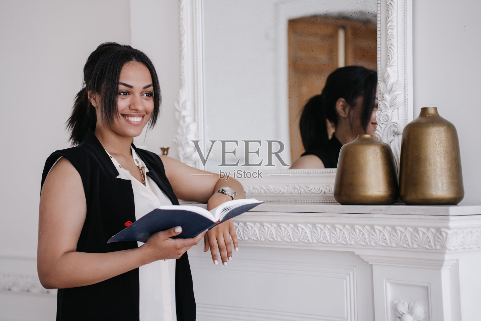 迷人的、快乐的非裔美国年轻女子靠在壁炉上，拿着日记，看向一边。美丽的巴西学生女孩在家里学习。年轻女企业家拿着笔记本站在家里。照片摄影图片