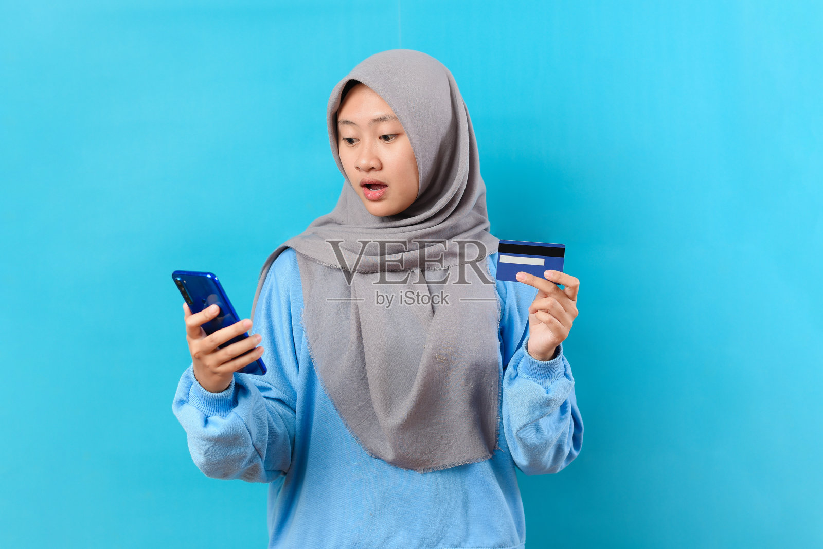 年轻的印尼妇女拿着信用卡，震惊地看着手机屏幕照片摄影图片