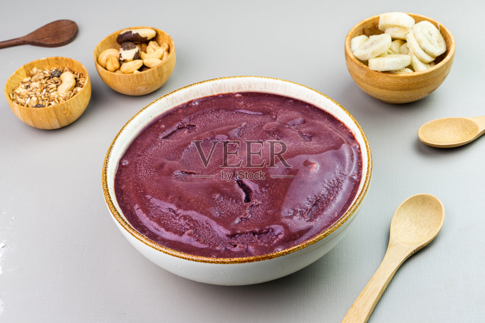 Brazilian açai in a white bowl, additional banana, granola and nuts in small bamboo bowls. Cashew and Pará nuts. Top view. Gray background. Selective focus照片摄影图片