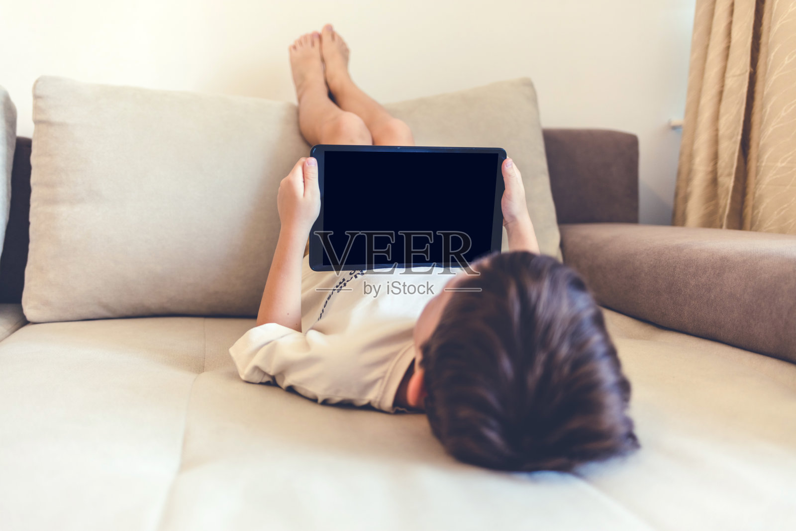 一个小男孩躺在沙发上使用他的平板电脑。男孩在家里用平板电脑玩或看东西。照片摄影图片