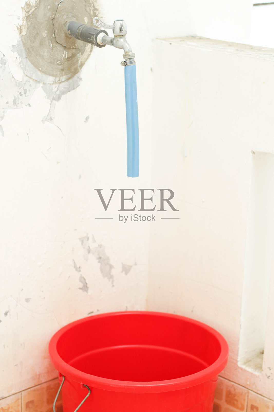 红色的塑料桶用来收集干净的水，上面是一个水龙头。红色的桶在房子的角落，对着瓷砖墙的背景。副本的空间。照片摄影图片