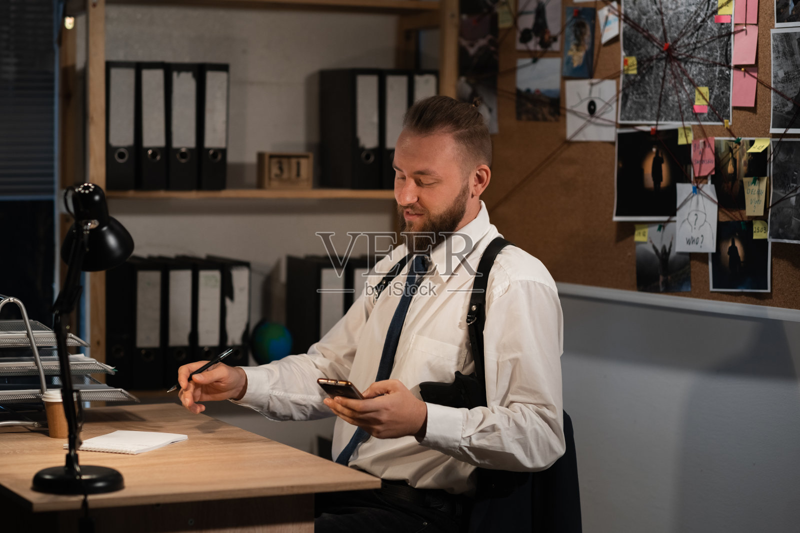 一位职业侦探晚上在办公室调查一起谋杀案。他坐在桌子旁，手里拿着智能手机，在网上阅读新闻。照片摄影图片