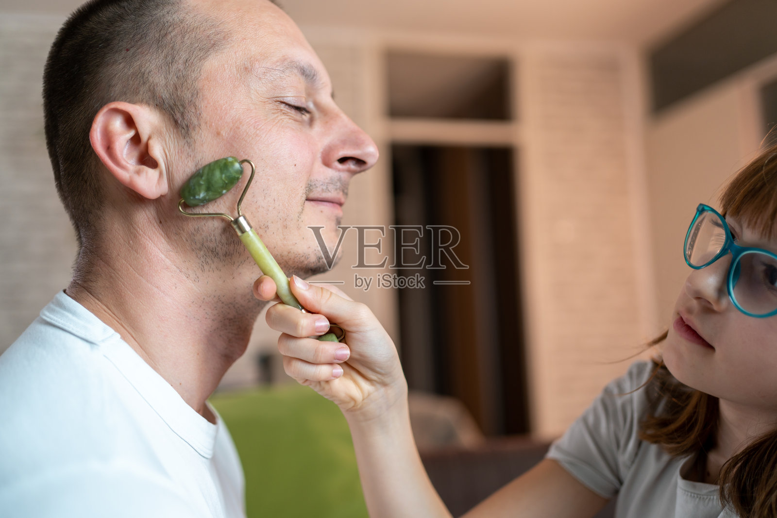 好奇的女儿使用面部按摩辊为她的父亲面部按摩。照片摄影图片