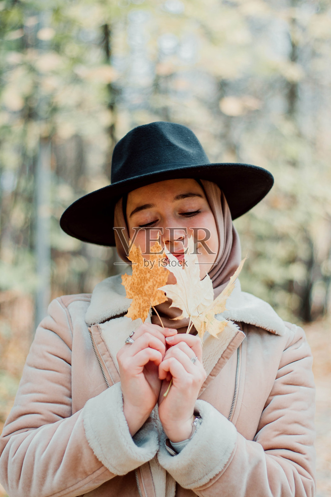 穿着米色外套和头巾的穆斯林妇女。时髦的穆斯林女人在公园里。一个戴着黑帽子的女孩坐在长凳上，手里拿着秋枫黄和橘色的叶子照片摄影图片