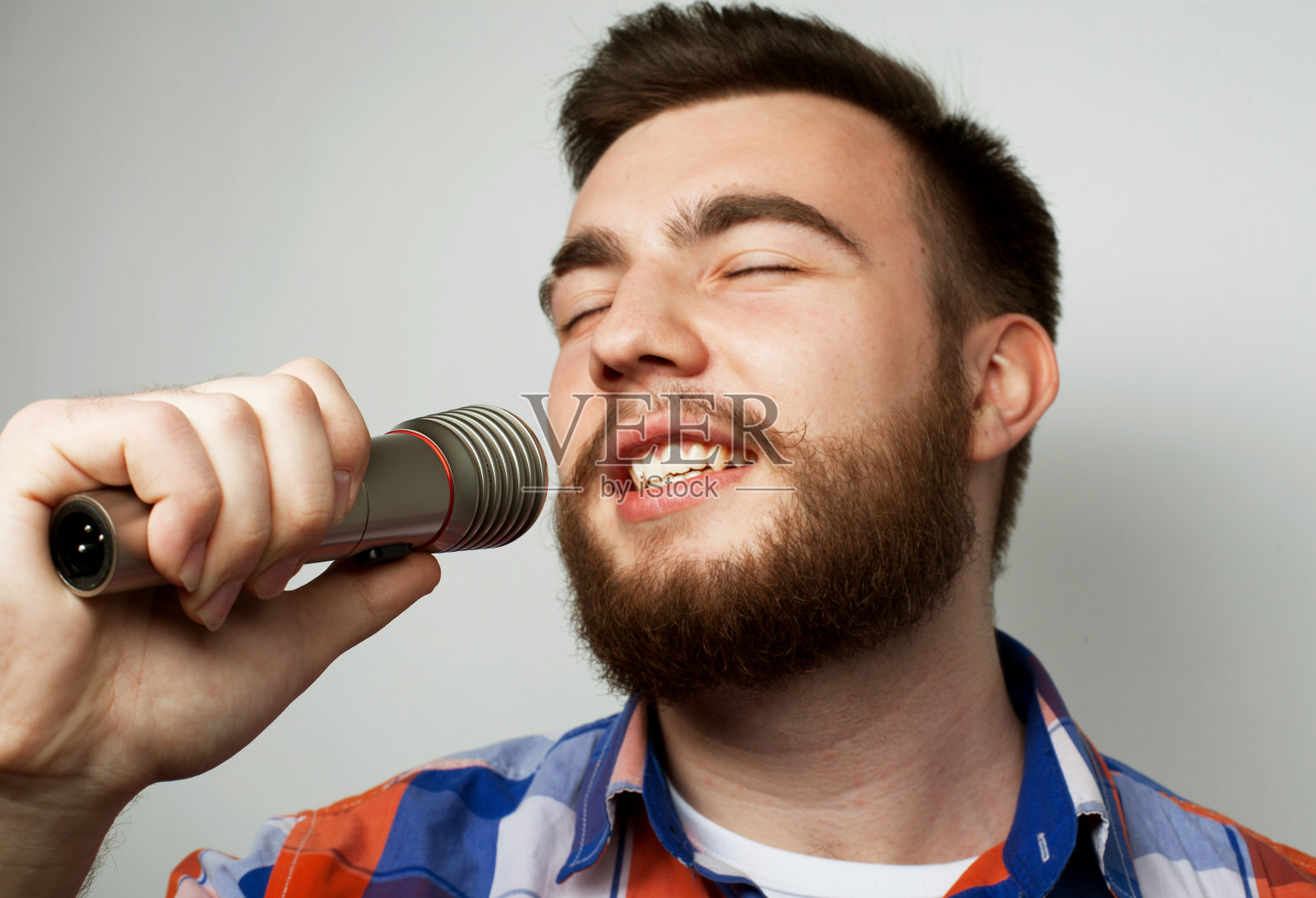 一个留着胡子、穿着格子衬衫的年轻人拿着麦克风唱歌。孤立在白色上。照片摄影图片