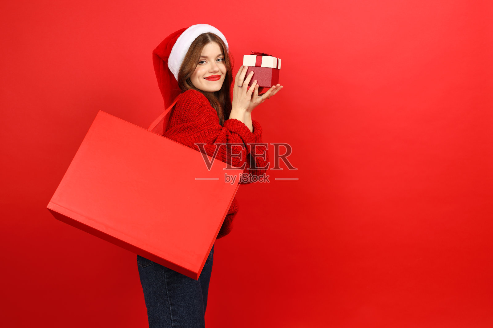 节日折扣和促销。快乐的有吸引力的女人与礼物和红色包装穿着圣诞老人的帽子和毛衣。照片摄影图片
