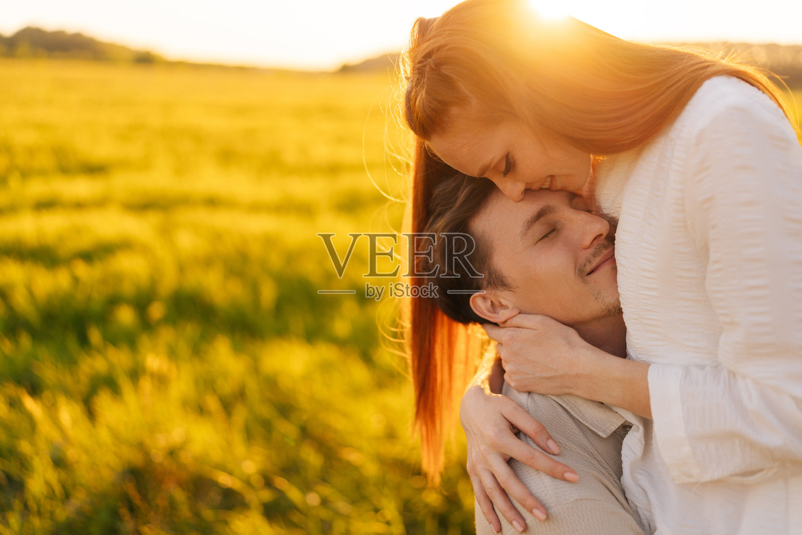 夏日黄昏，金色夕阳，温暖阳光，一对相爱的年轻夫妇闭着眼睛站在美丽的绿色草地上温柔地拥抱。照片摄影图片