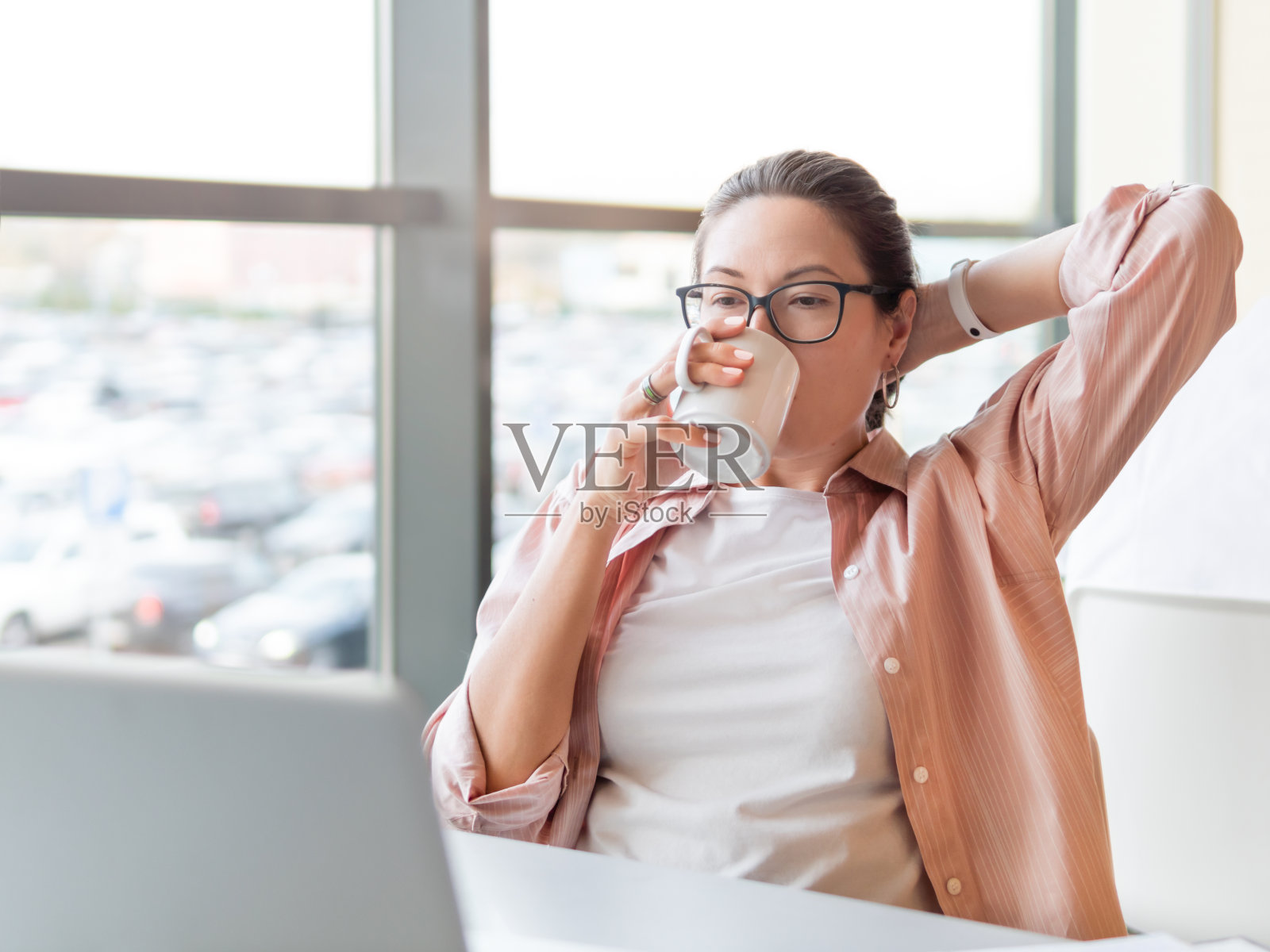 放松的女人喝咖啡，用笔记本电脑工作。在联合办公中心设有全景窗户的现代办公室。商务中心自由职业者或学生的工作场所。照片摄影图片