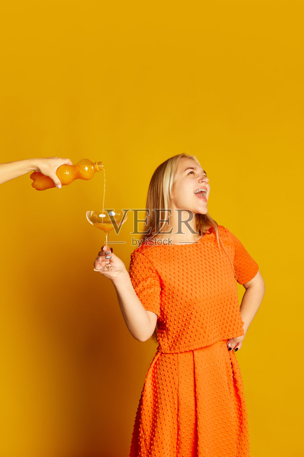派对时间。激动年轻美丽的女孩在橙色礼服品尝鸡尾酒，有乐趣孤立在橙色背景。生动的情感，快乐，爱好和快乐照片摄影图片