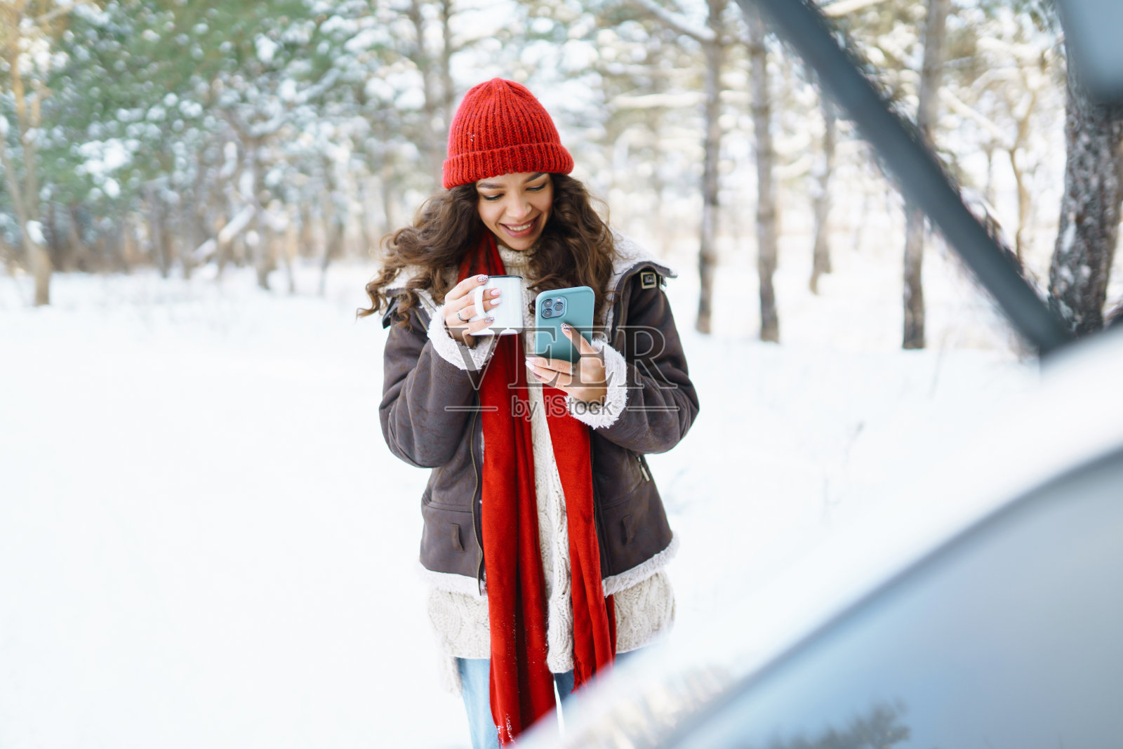 在冬天的森林里，快乐的女人拿着保温杯坐在汽车后备箱里喝茶。照片摄影图片