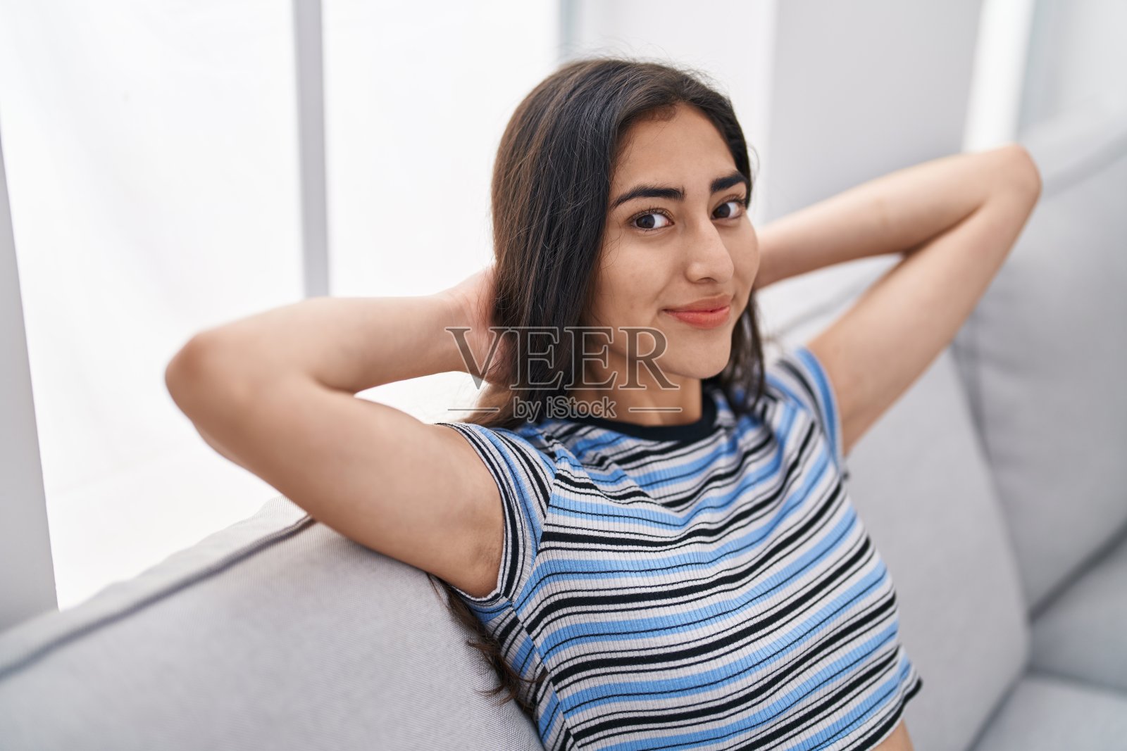 年轻的西班牙女孩手放在头上放松坐在家里的沙发上照片摄影图片