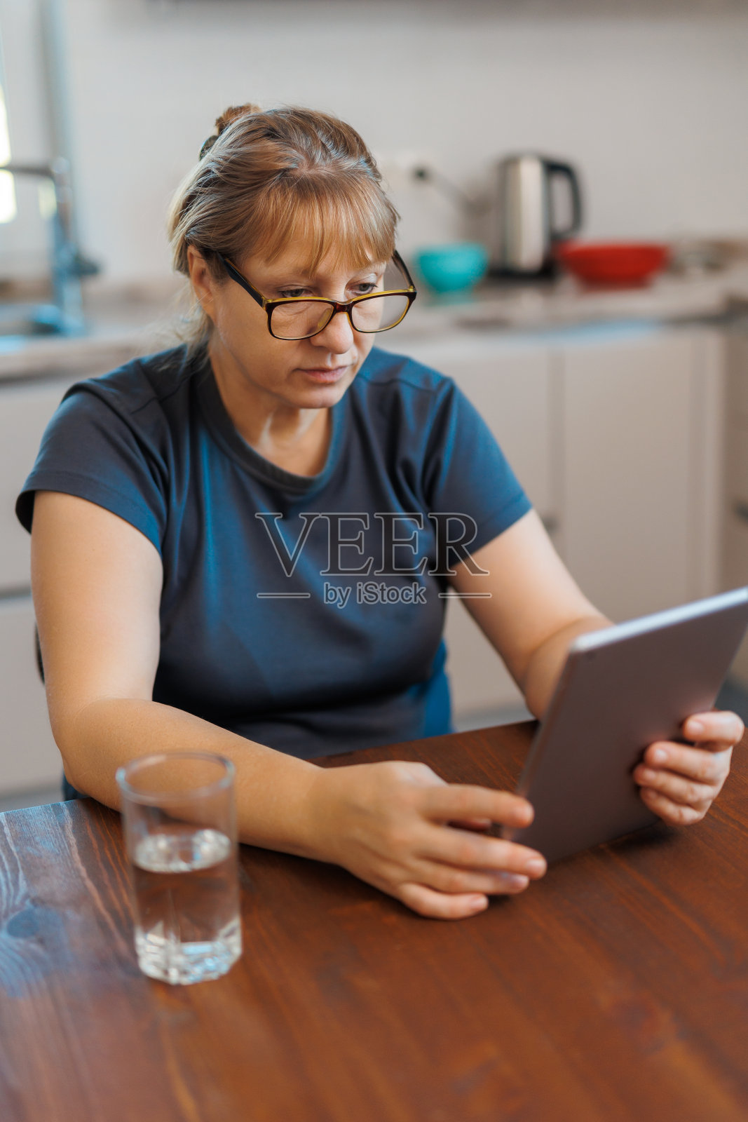 快乐的中年50岁妇女使用数字平板电脑坐在家里的厨房。成熟的老太太戴眼镜抱着平板电脑看电子书，网上购物，打视频电话。照片摄影图片