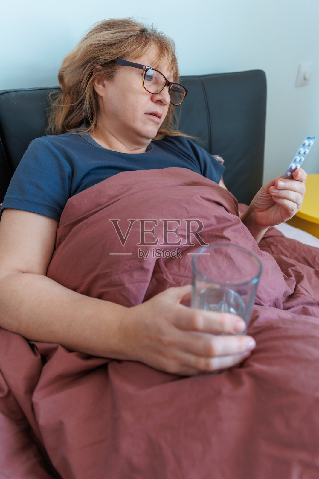 生病的老妇人躺在家里的床上感到生病，患流感或感冒用处方药治疗。女人用杯子喝水照片摄影图片