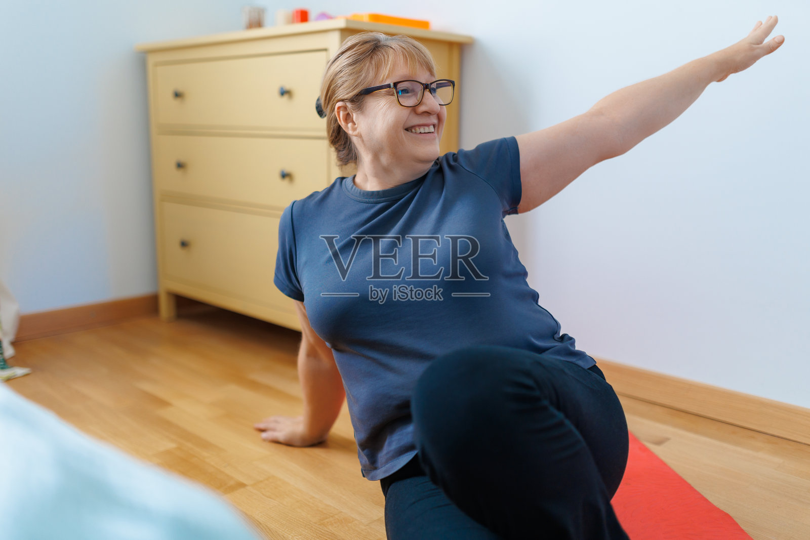 活跃的老年妇女在家里的瑜伽垫上做腹肌练习，对着镜头微笑。运动成熟的女士在客厅内部工作。力量训练概念照片摄影图片