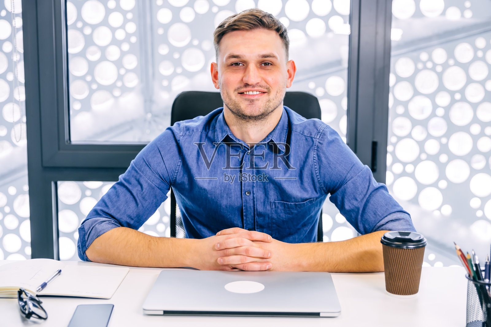 一个英俊的微笑的男人在办公室里做一个项目，看着相机，把笔记本电脑放在面前，一边喝着咖啡，一边写着想法。照片摄影图片