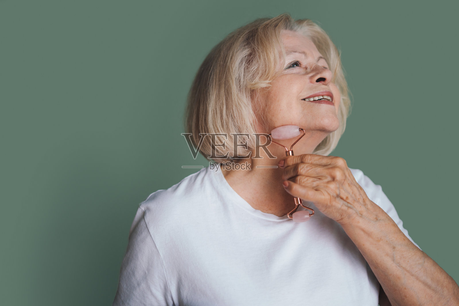 可爱的老年妇女做玉石轮面部按摩刺激淋巴引流，孤立的绿色背景。Spa技术护肤广告。抗衰老美容理念。照片摄影图片