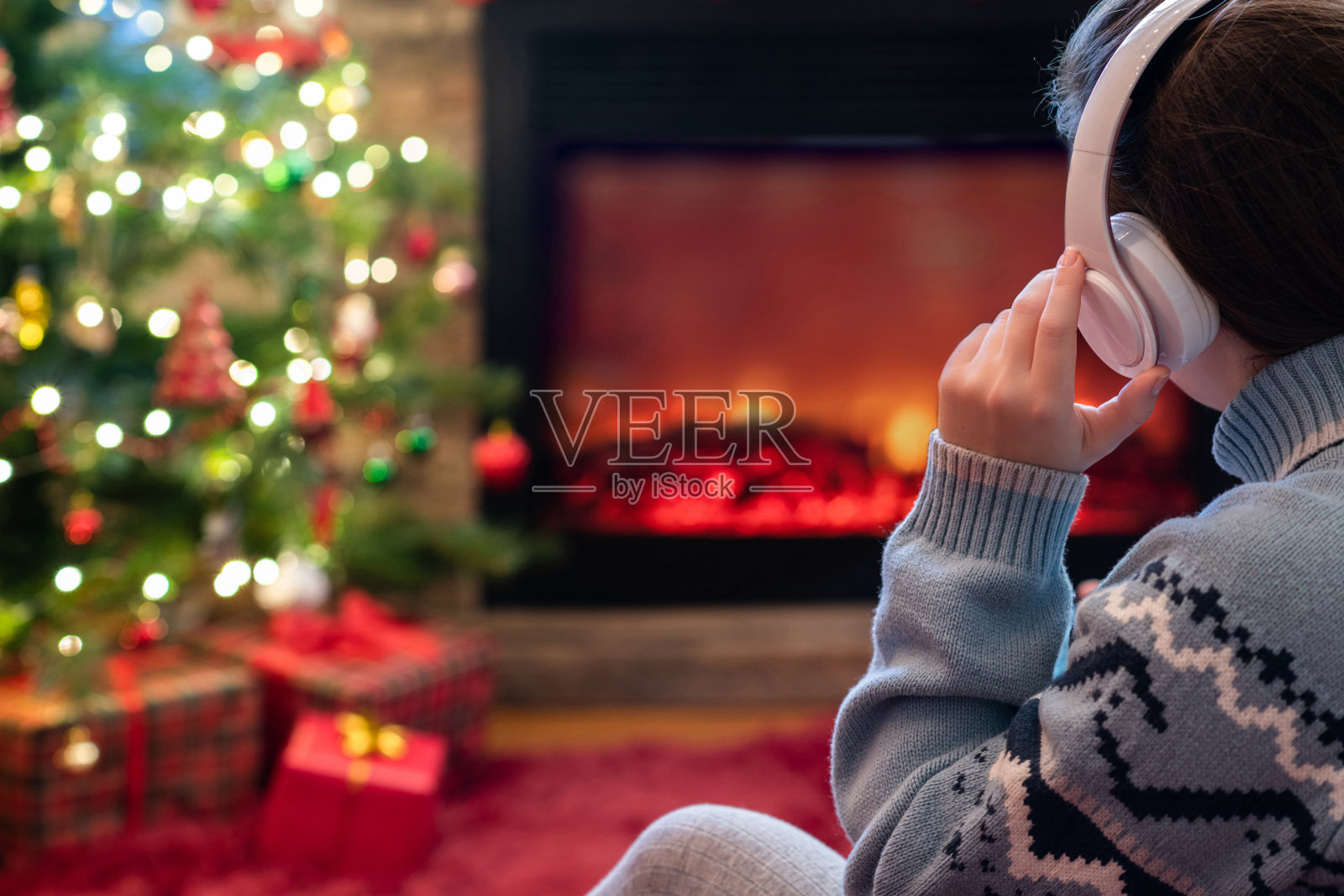 在冬天的晚上，戴着耳机的女人坐在壁炉火焰和圣诞树旁取暖。照片摄影图片
