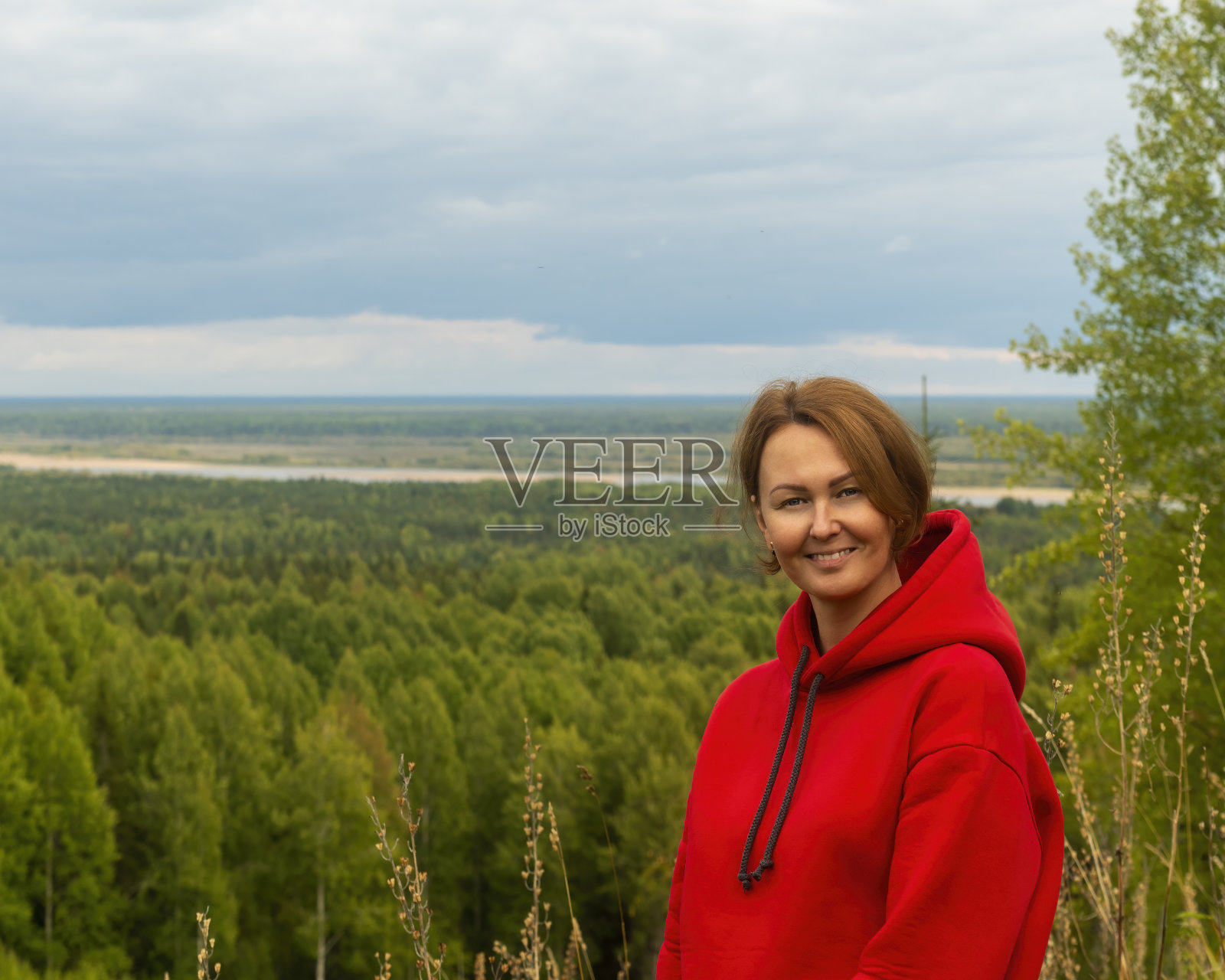 一个红头发和红色卫衣的女人站在树木繁茂的小山上，背景是森林和远处蜿蜒的河流。照片摄影图片