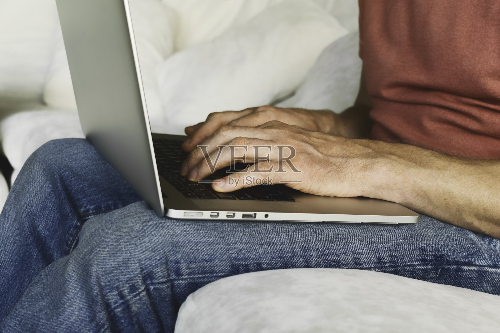 一个穿着牛仔裤和t恤的男人坐在沙发上，在笔记本电脑上打印。男人的手，在电脑上打印，特写照片摄影图片