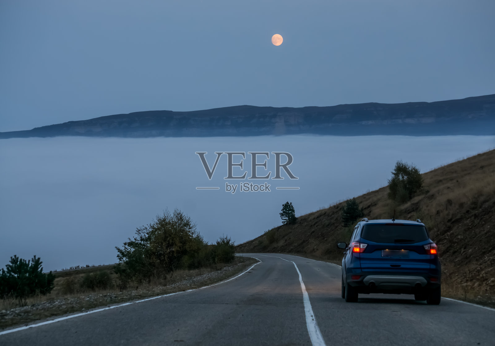 一辆蓝色的汽车行驶在山路上，在满月的背景下，在山谷的云层中下降。初秋的早晨，橙色的月亮在山上照片摄影图片