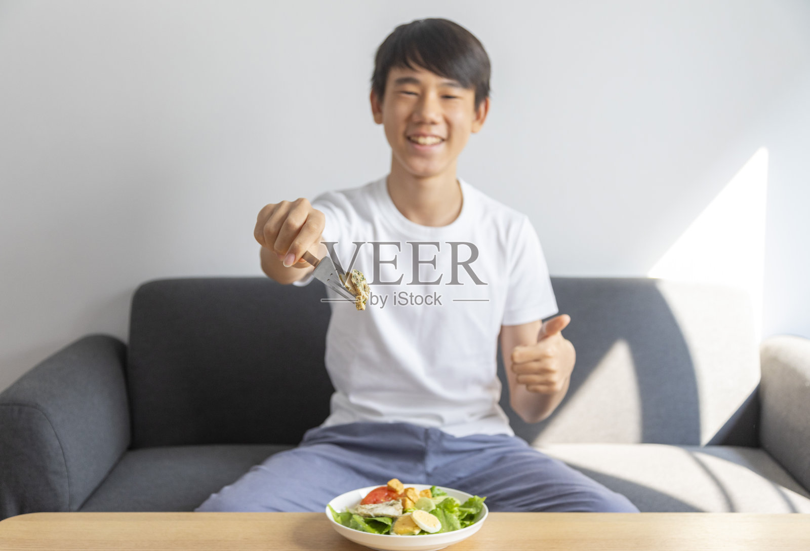 亚洲十几岁的男孩微笑着开心地端着一碗新鲜的沙拉，坐在房间的沙发上。照片摄影图片