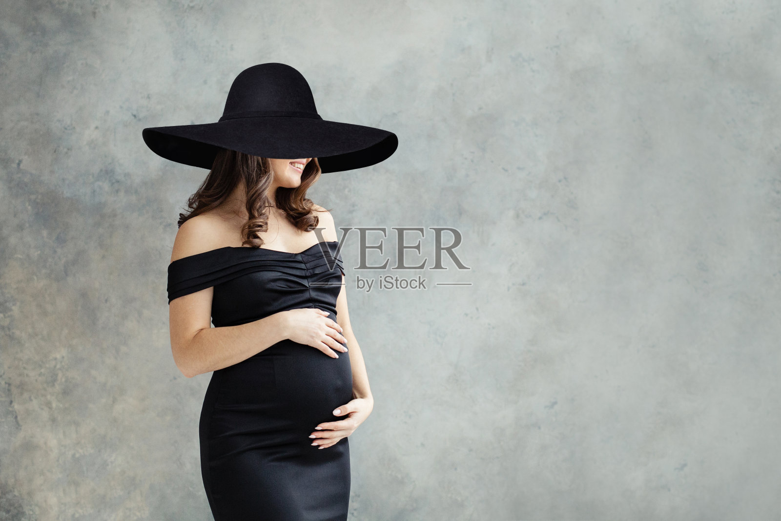 开朗的孕妇在宽黑色宽边帽肖像。美丽的女士黑发穿着黑色礼服广告微笑在灰色的工作室背景照片摄影图片