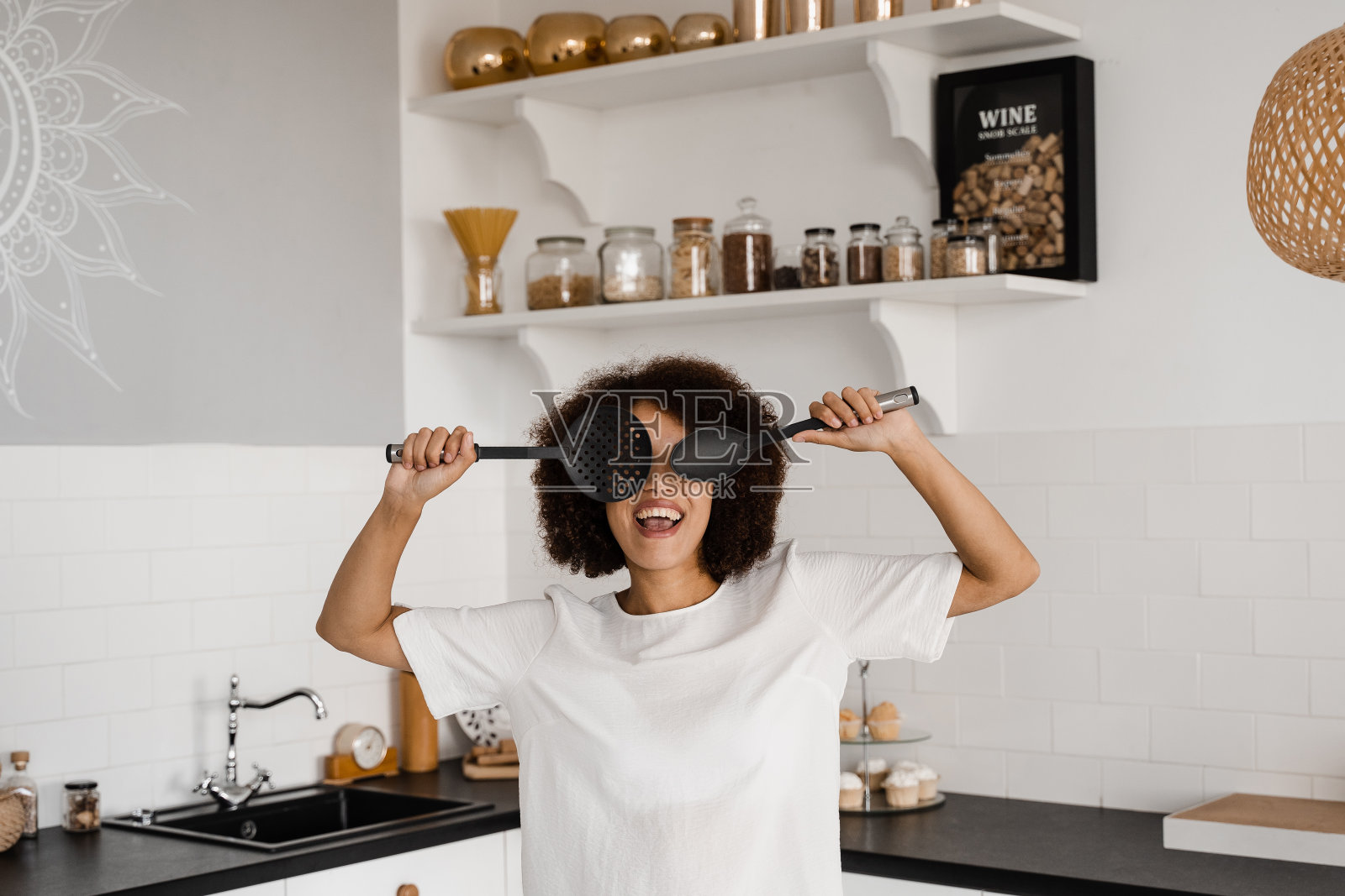 身穿围裙的非洲家庭主妇在厨房里拿着做饭用的锅铲摆姿势。非裔美国女孩厨师在厨房玩得很开心。烹饪课程和美食工作坊的广告。照片摄影图片