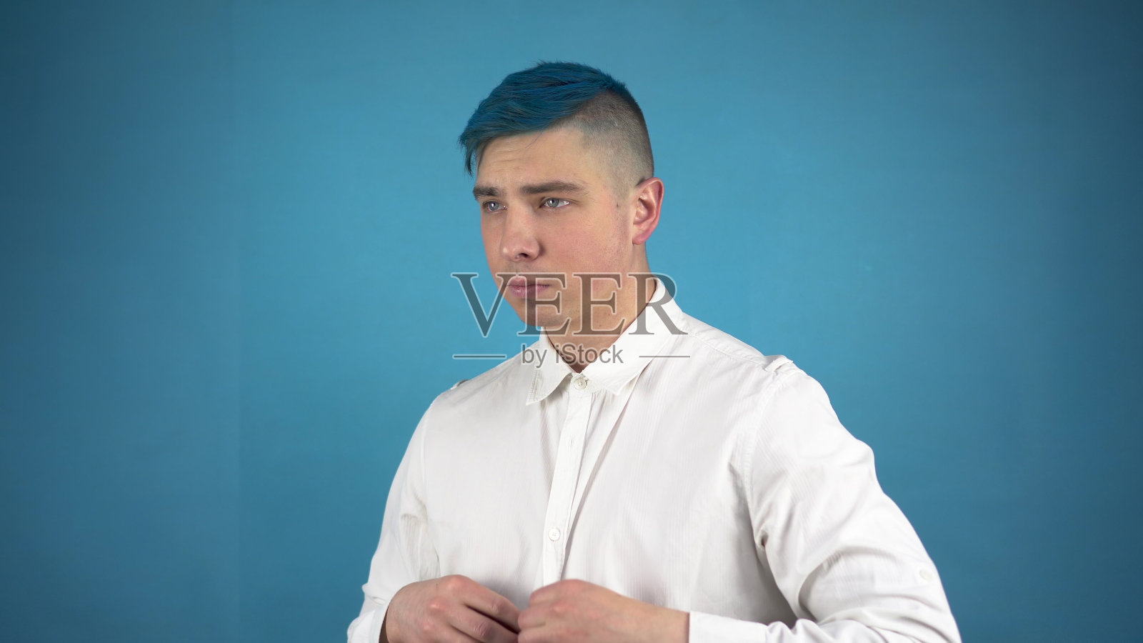 一个戴眼镜、蓝头发的年轻人正在打扮西装。另一个人系上扣子，在蓝色背景上给自己系上领带。照片摄影图片