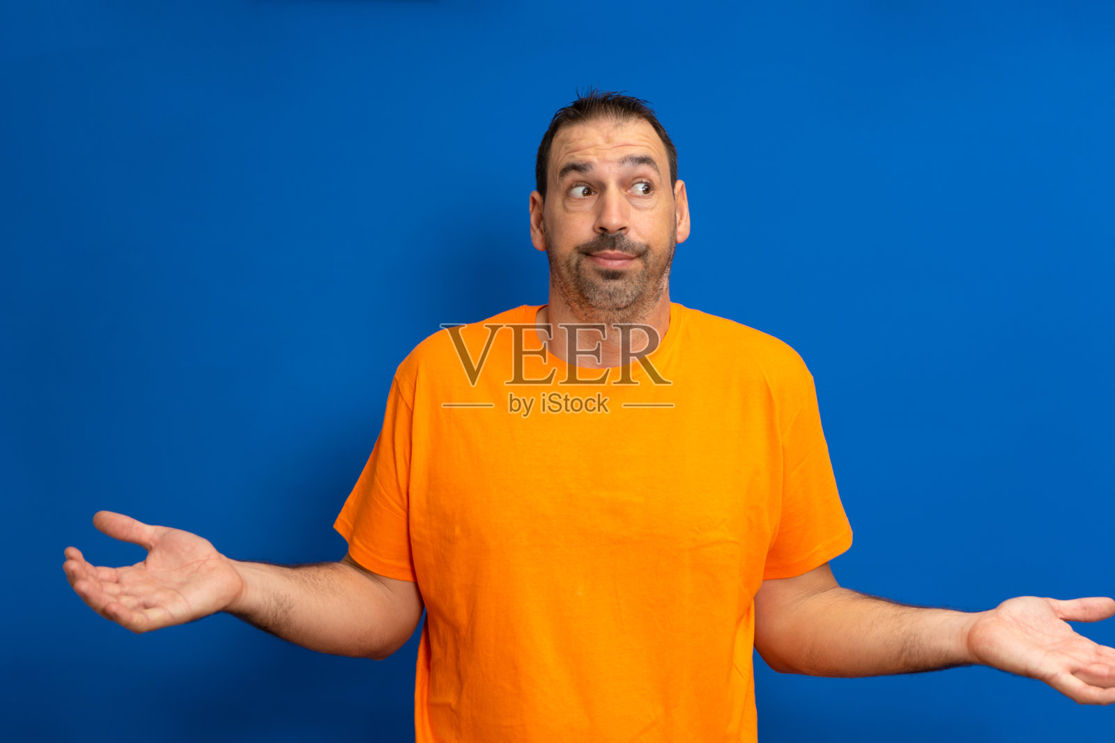 穿着橙色t恤的拉丁大胡子男子孤立在蓝色背景怀疑和耸肩的质疑姿态照片摄影图片